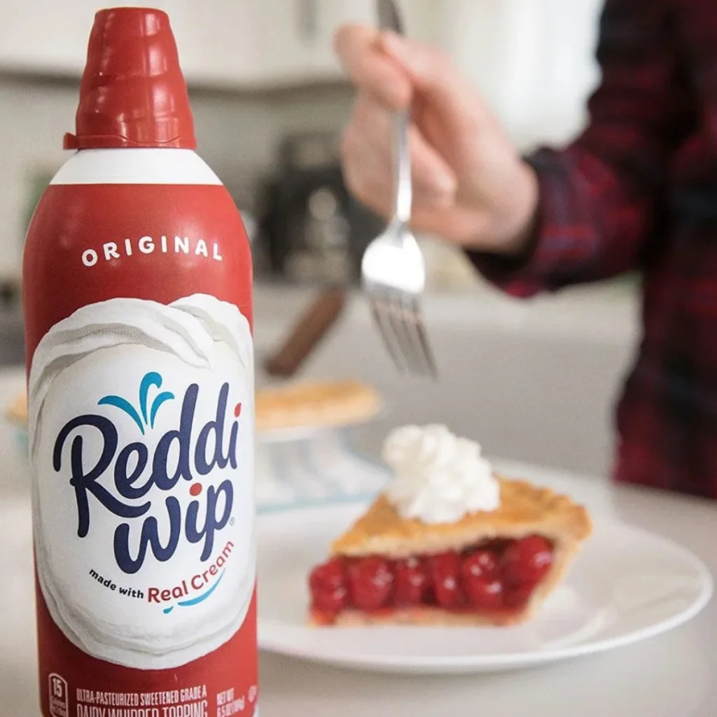 A can of Reddi Wip whipped cream in the foreground, with a slice of cherry pie topped with whipped cream on a plate in the background. A hand holding a fork is reaching towards the pie.